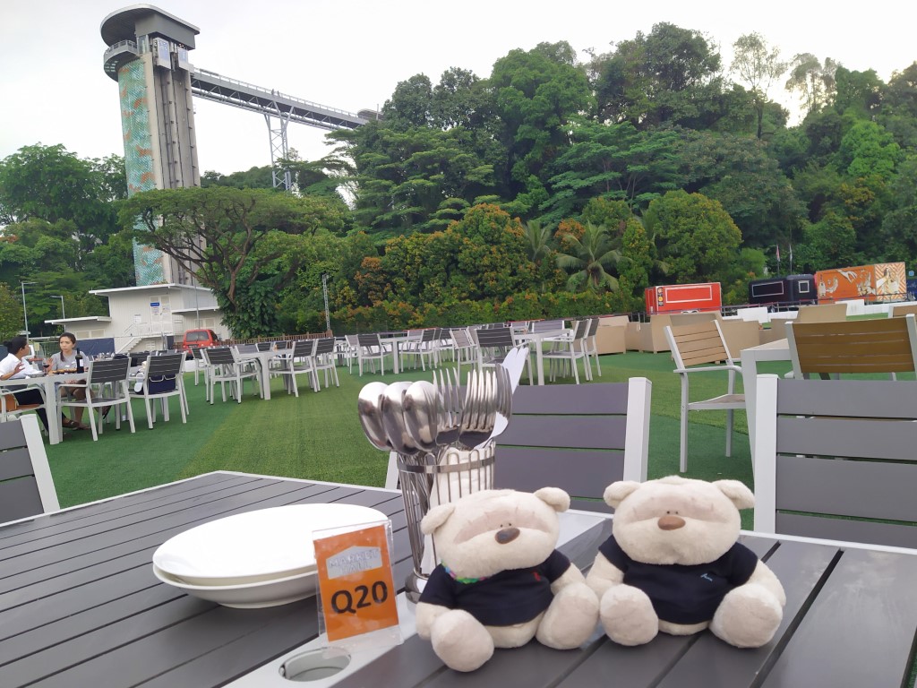 2bearbear seated outdoors at Market Hall Sentosa with Fort Siloso Skywalk in the background